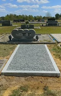Gravestone with surround, engraved headstone featuring four heart shapes in a cemetery with other graves and trees in the background.