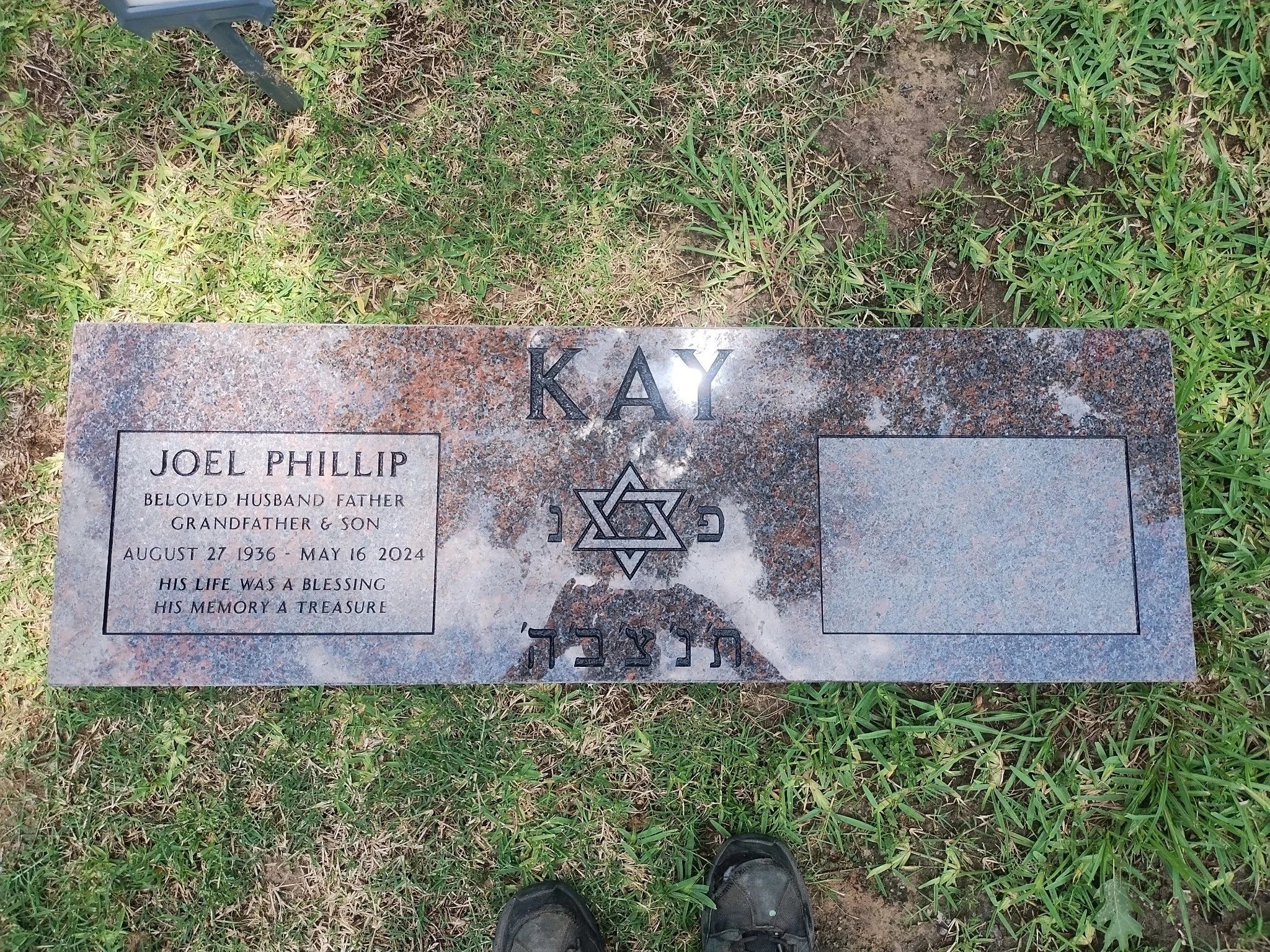 A gravestone on the ground surrounded by grass. The gravestone has the name 'KAY' engraved at the top, with a Star of David symbol and Hebrew inscriptions below. The lower part of the gravestone contains the name 'Joel Phillip,' born August 27, 1936, and died May 16, 2024, with a quote about his life and memory.