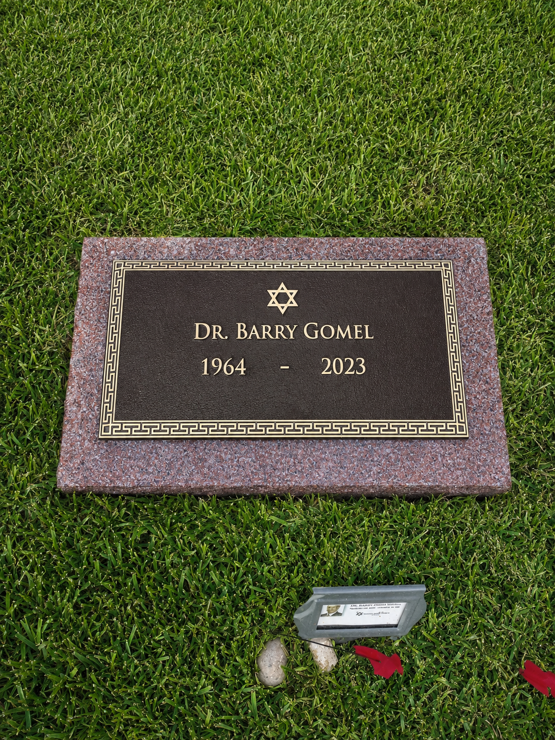 Gravestone of Dr. Barry Gomel, 1964-2023, with Star of David symbol, set on a stone base, surrounded by green grass and a small memorial plaque at the front.
