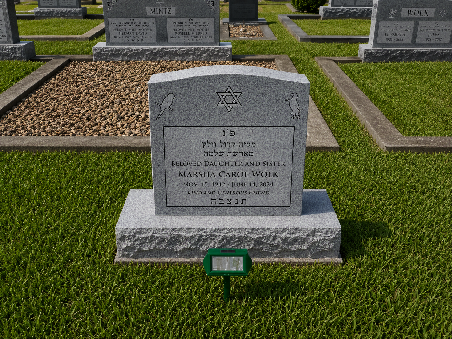 Gravestone in a cemetery with Hebrew and English inscriptions, commemorating Marsha Carol Wolk (November 15, 1942 - June 14, 2024), described as a beloved daughter, sister, and friend, with symbols of a star of David, a bird, and a rabbit or dog engraved on top.