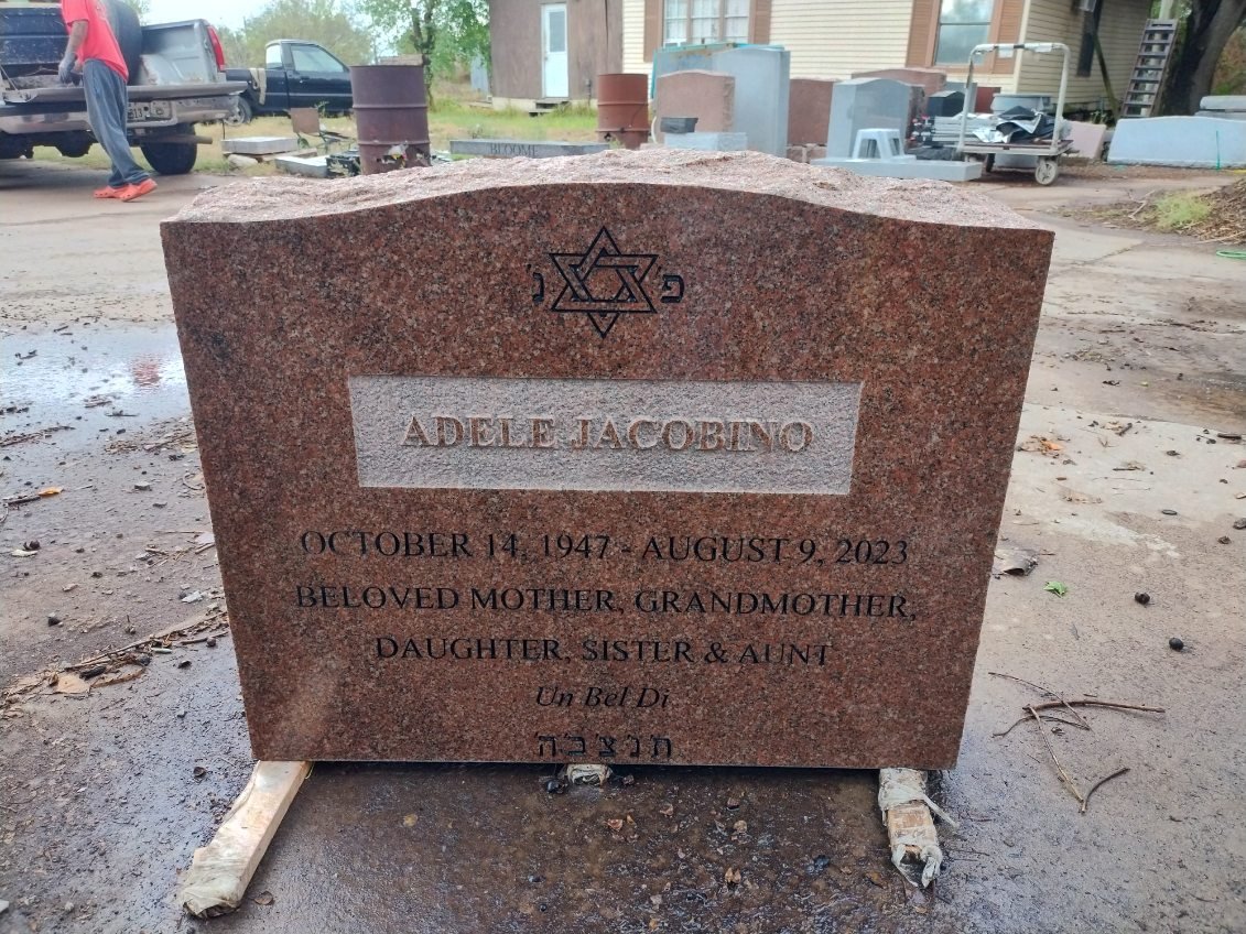 Gravestone with Star of David at the top, located outdoors in a cemetery or memorial site, with tools and other stones in the background.