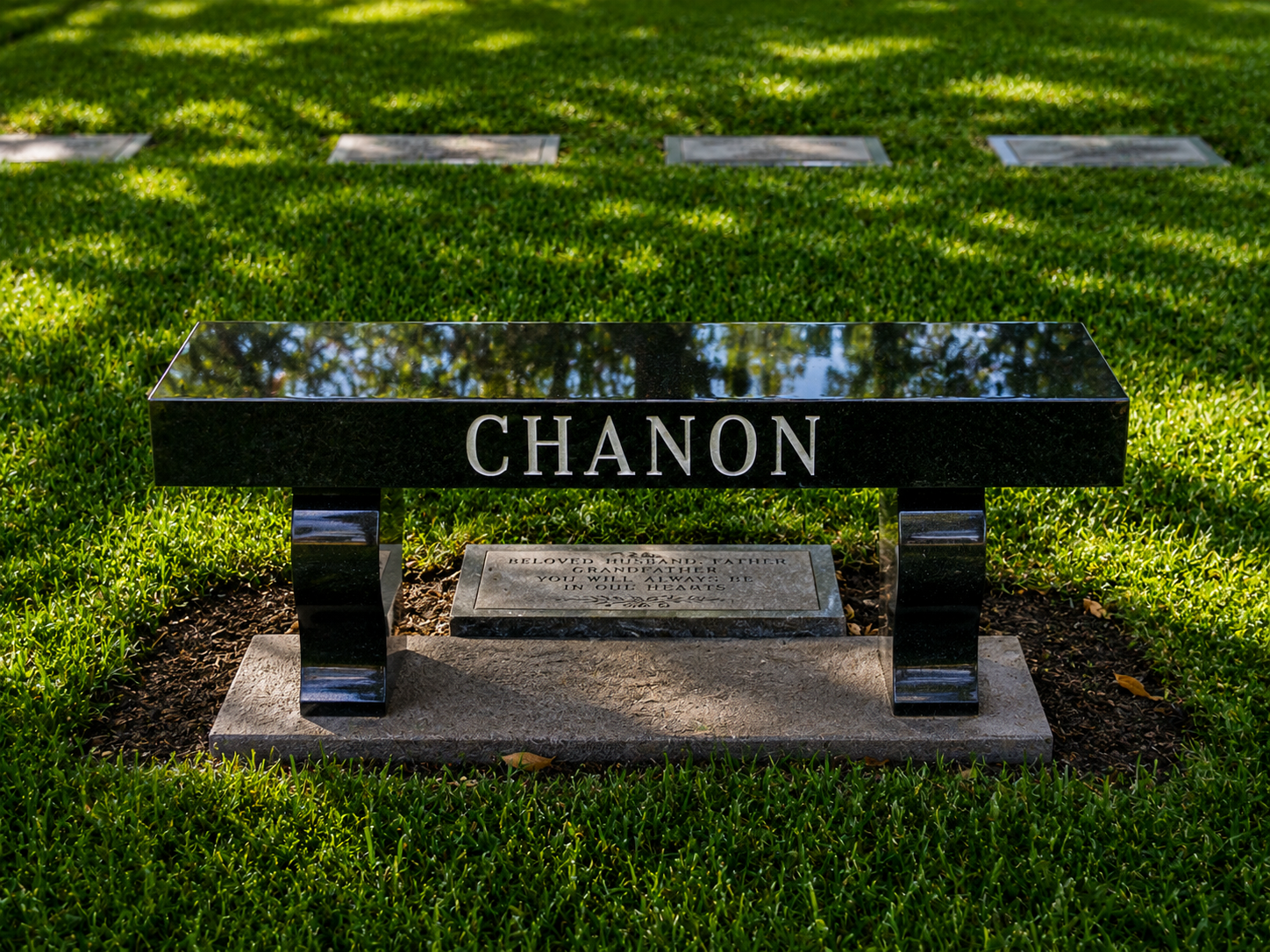 Gravestone with the name 'Chanon' in a cemetery, surrounded by green grass, with a reflective black bench.
