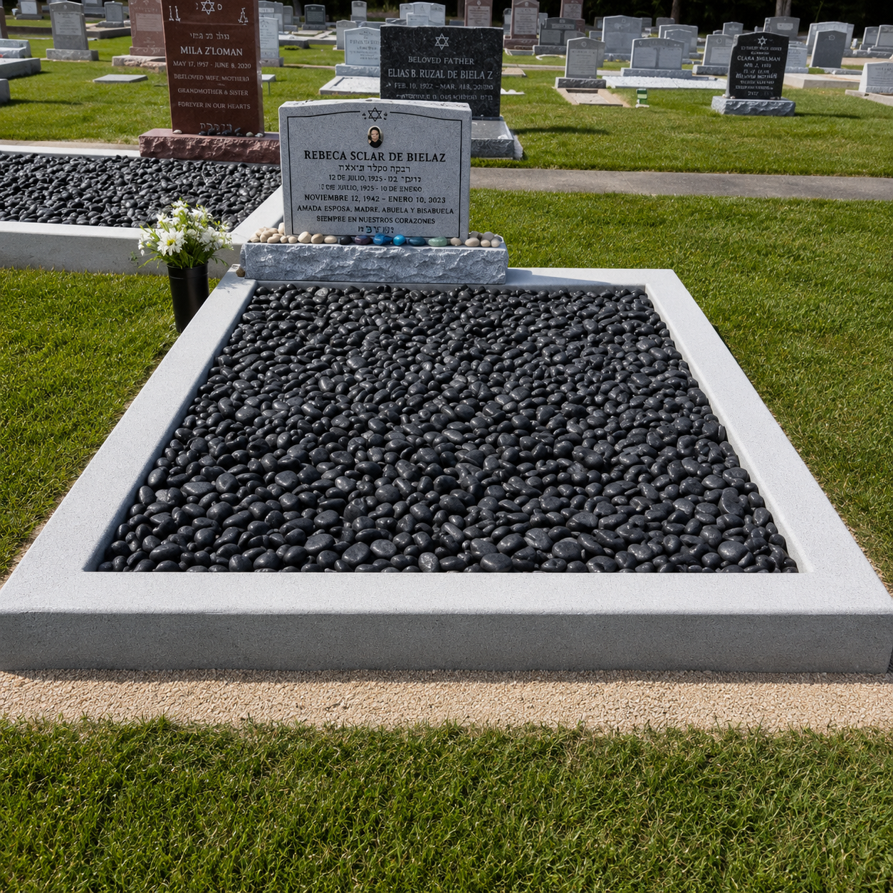 Gravestone in a cemetery with black pebbles surrounding the headstone, which has an inscription in Spanish and a small photo of the deceased. There is a small black vase with white flowers next to the headstone.