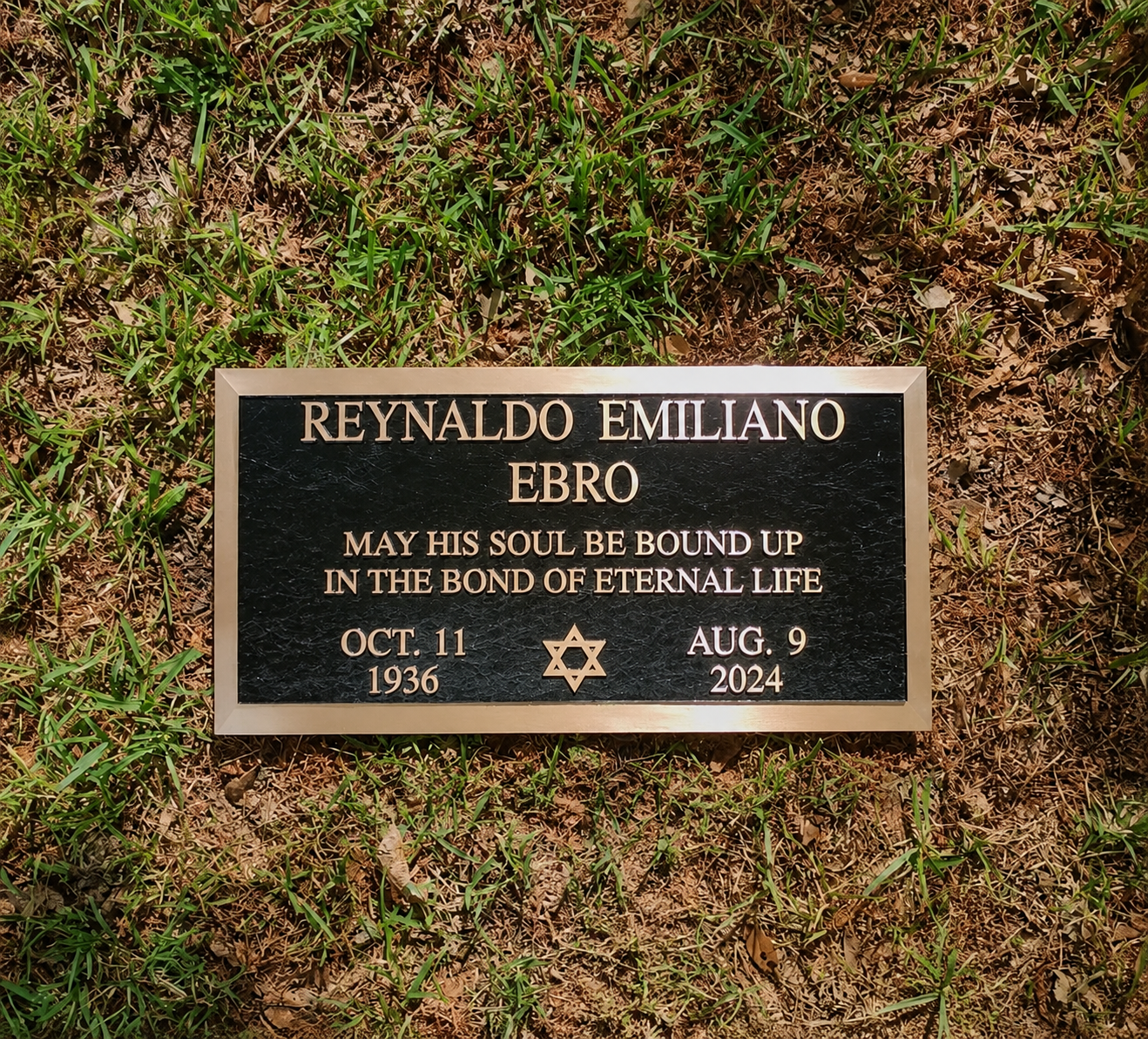 Gravestone with black plaque surrounded by grass, inscribed with the name Reynaldo Emiliano Ebro, with dates October 11, 1936, and August 9, 2024, and a Star of David symbol.