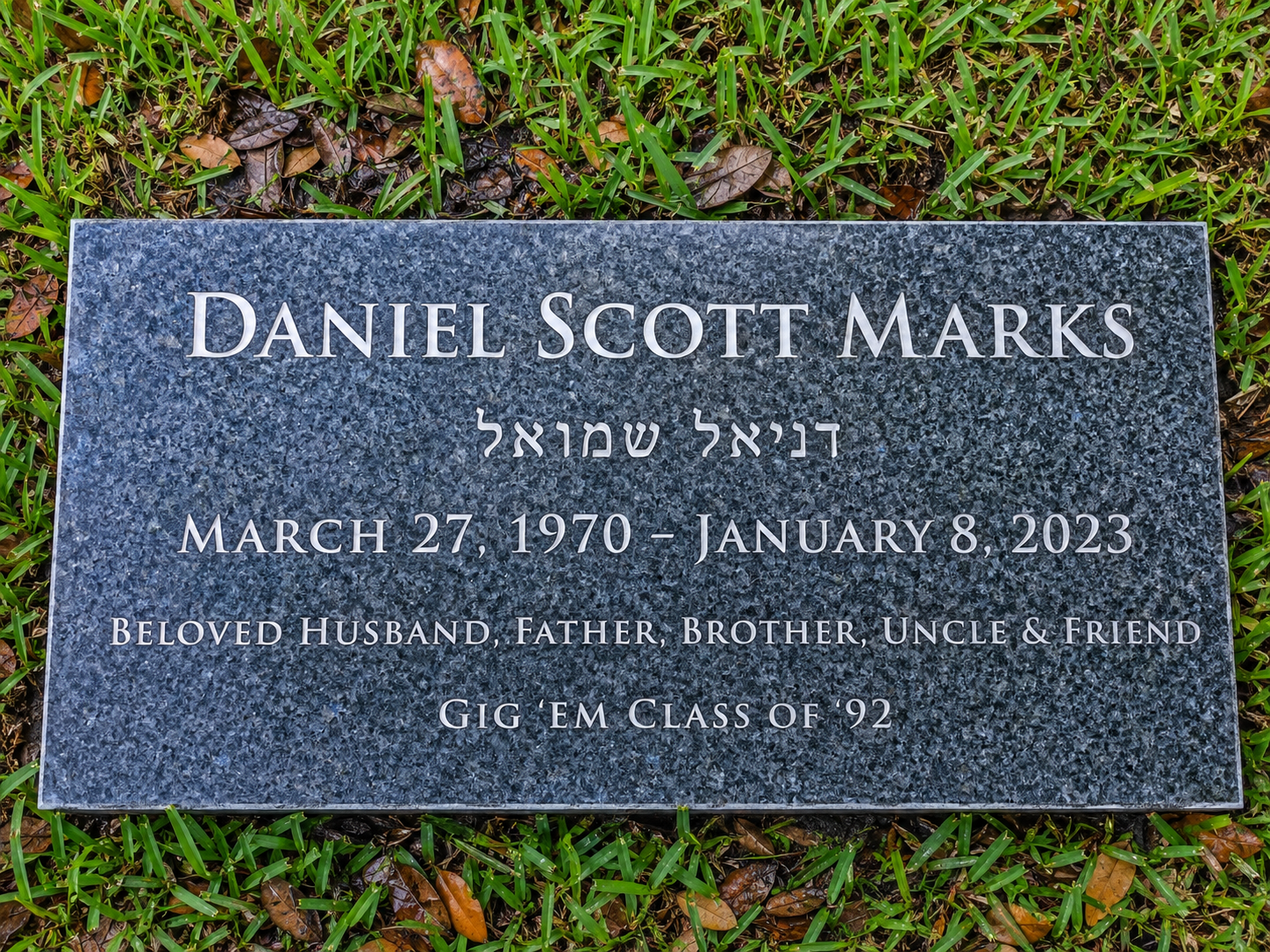 Gravestone plaque for Daniel Scott Marks, born March 27, 1970, died January 8, 2023, surrounded by green grass and fallen leaves.