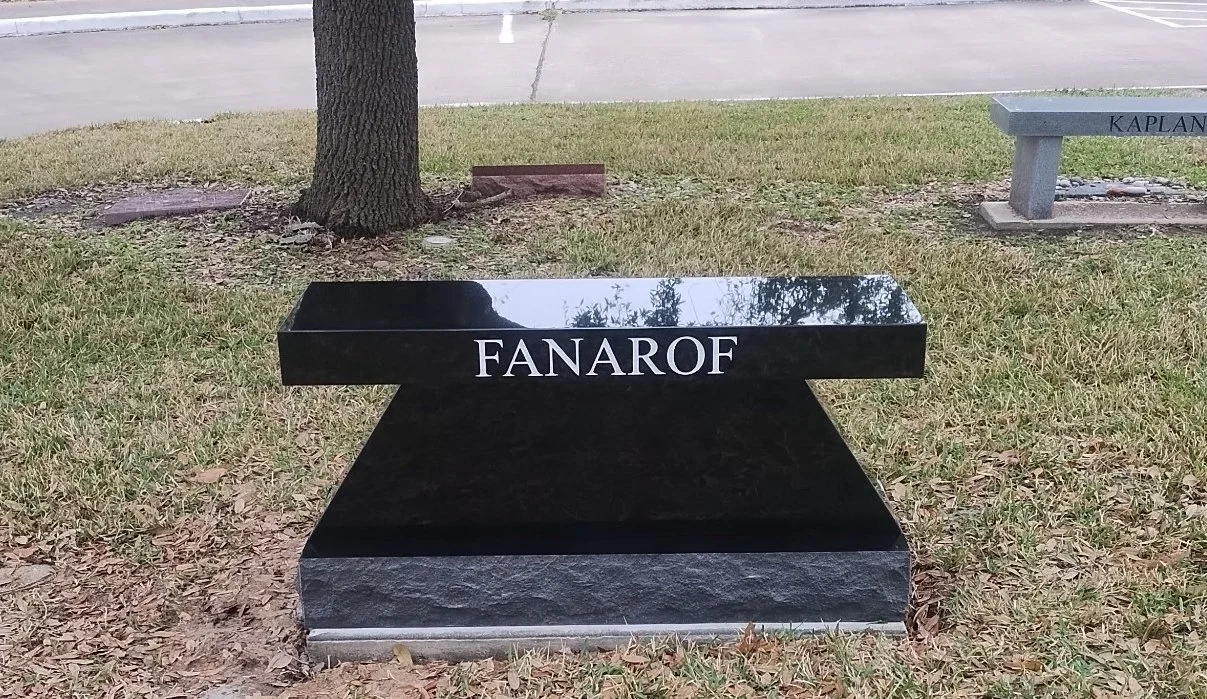 Black granite memorial bench engraved on the backrest, situated on a grassy area near a tree and a paved road.
