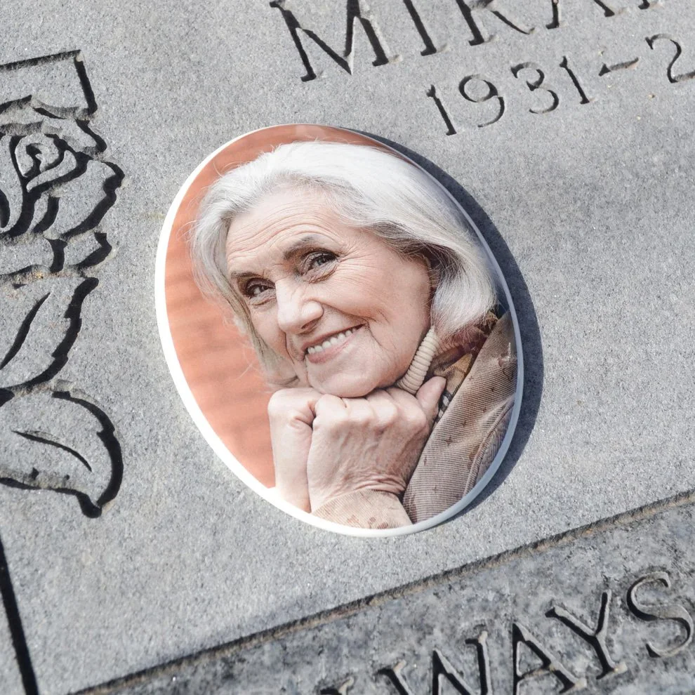 A close-up photograph of a woman with gray hair smiling and resting her chin on her hand, displayed in an oval-shaped frame embedded in a gray granite memorial or gravestone.
