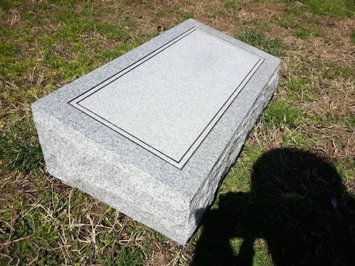 A gray granite gravestone resting on grass and dirt, with a blank rectangular engraved area.
