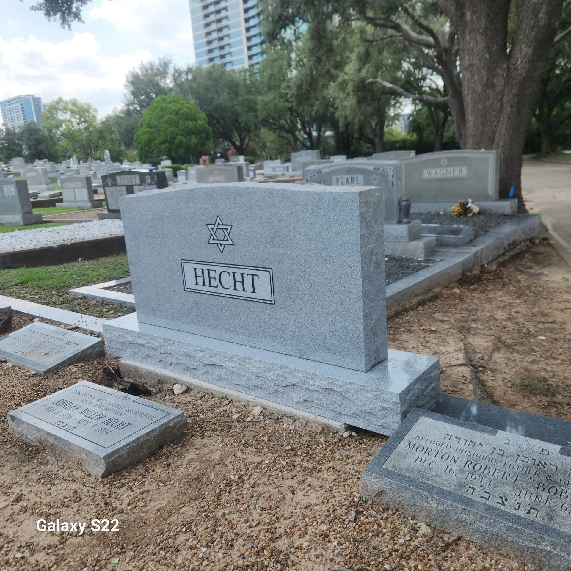 Gravestone in a cemetery with engraved Star of David, surrounded by other graves, trees, and city buildings in the background.
