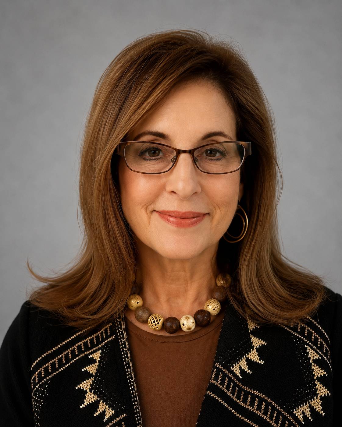 A woman with shoulder-length light brown hair wearing glasses, hoop earrings, a beaded necklace, and a black and beige embroidered jacket.