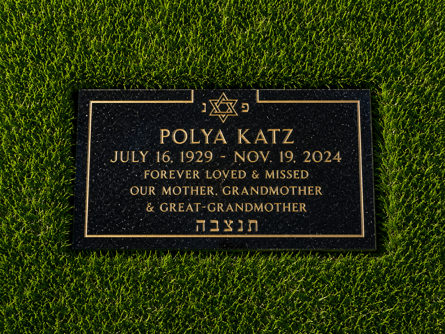 A black grave marker surrounded by green grass. The marker has gold Hebrew and English inscriptions, including a star of David at the top and the name 'Polya Katz' with dates July 16, 1929, to Nov. 19, 2024, and a message of love and remembrance.