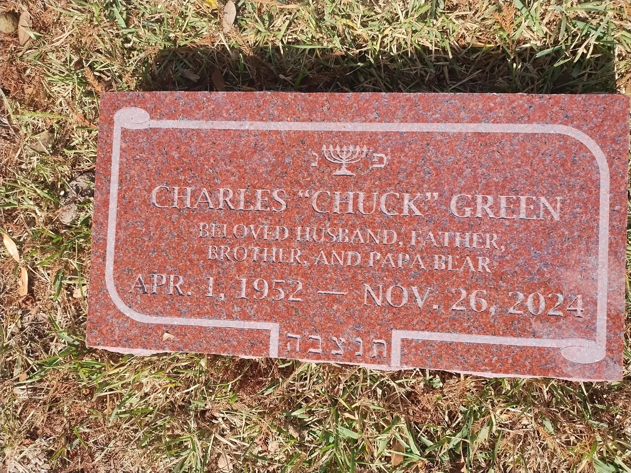 A memorial headstone made of red granite with carved out text. The headstone is dedicated to Charles 'Chuck' Green and mentions his birth date, April 1, 1952, and his death date, November 26, 2021. The headstone is placed on grass and surrounded by small plants.