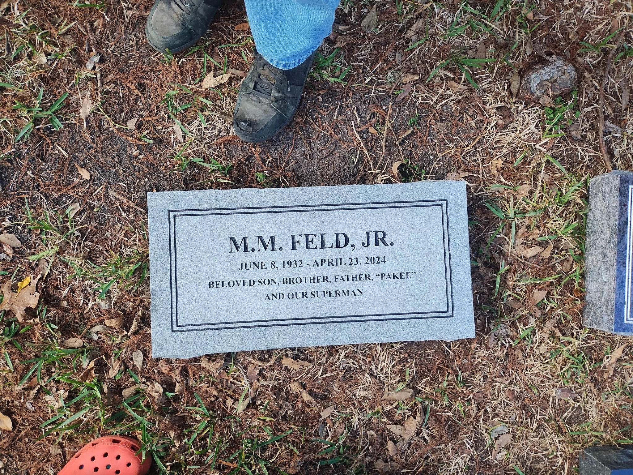 Close-up of a gravestone with inscription for M.M. Feld Jr., born June 8, 1932, died April 23, 2024. The gravestone is surrounded by grass, leaves, and small plants. Two shoes, one black and one gray, and an orange Croc are visible near the gravestone.