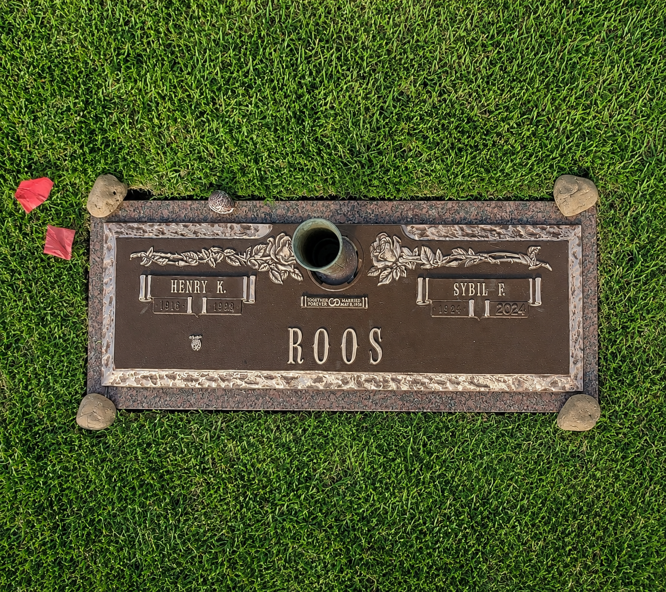 Gravesite plaque on the grass for Henry K. and Sybil F. Roos, with a candle and flowers nearby.