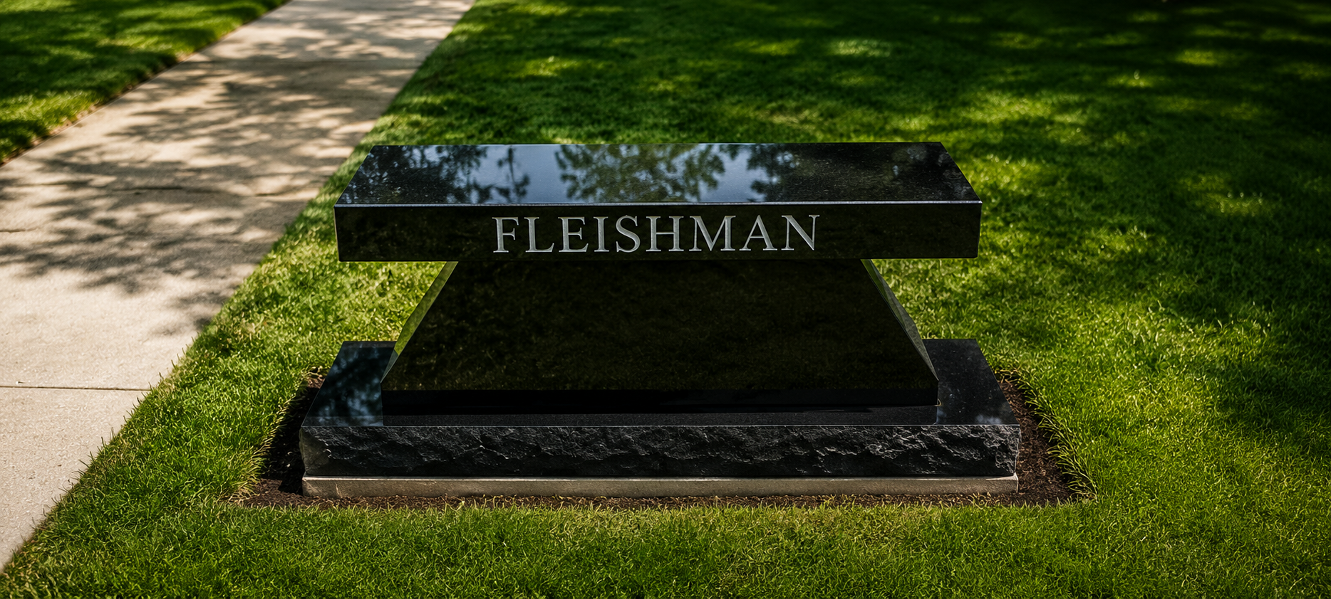 A black granite headstone with the name 'FLEISHMAN' engraved on it, located on a grassy area with a concrete sidewalk nearby.