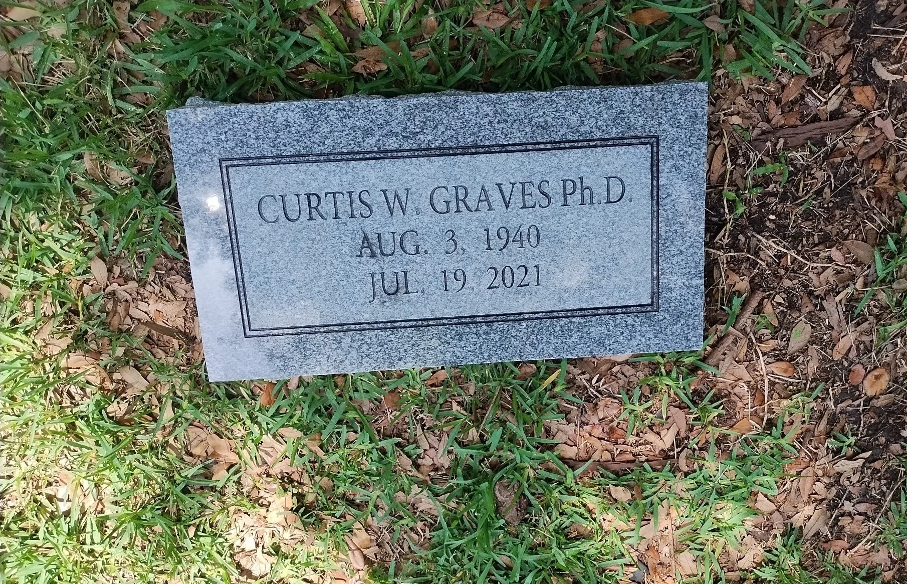 Gravestone with inscriptions for Curtis W. Graves Ph.D., born August 3, 1940, died July 19, 2021, lying among grass and fallen leaves.