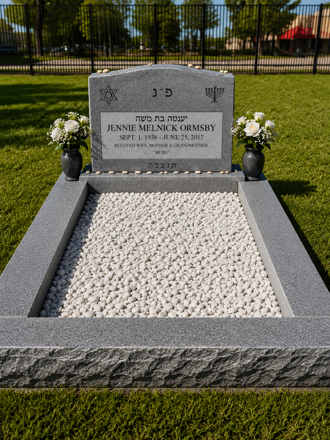 Gravestone for Jennie Melnick Ormsby, with white flowers in black vases on each side, white pebbles inside the grave, and a black fence in the background.