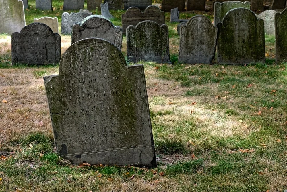 A weathered gravestone in a cemetery with other gravestones visible in the background.