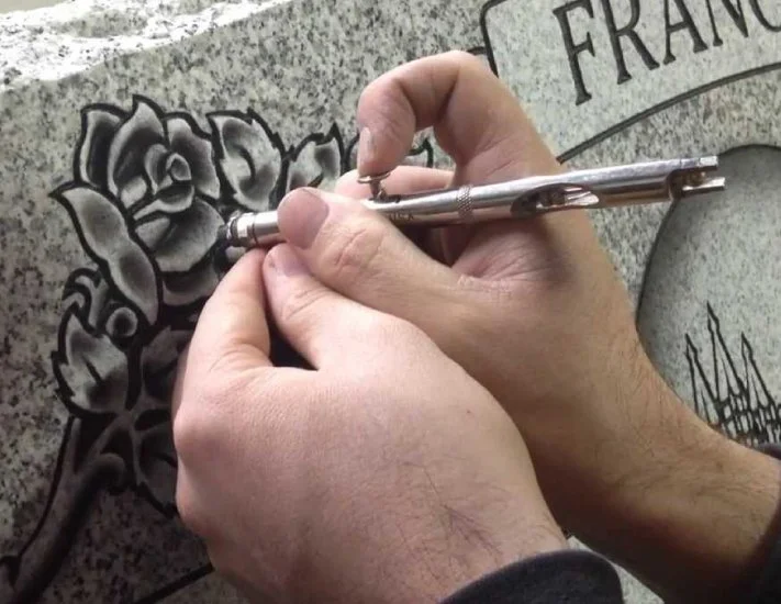 Person's hands inscribing a floral design on a stone memorial or gravestone. The stone has rounded top edges with text visible, including the partial word 'FRANCE'.