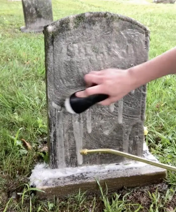 Person scrubbing a gray stone headstone with a handheld brush, as water and soap drip down the stone, in a grassy area of a cemetery.