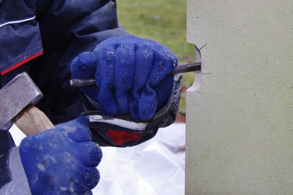A person in blue gloves is using a chisel and hammer to carve a hole into a light-colored concrete wall.