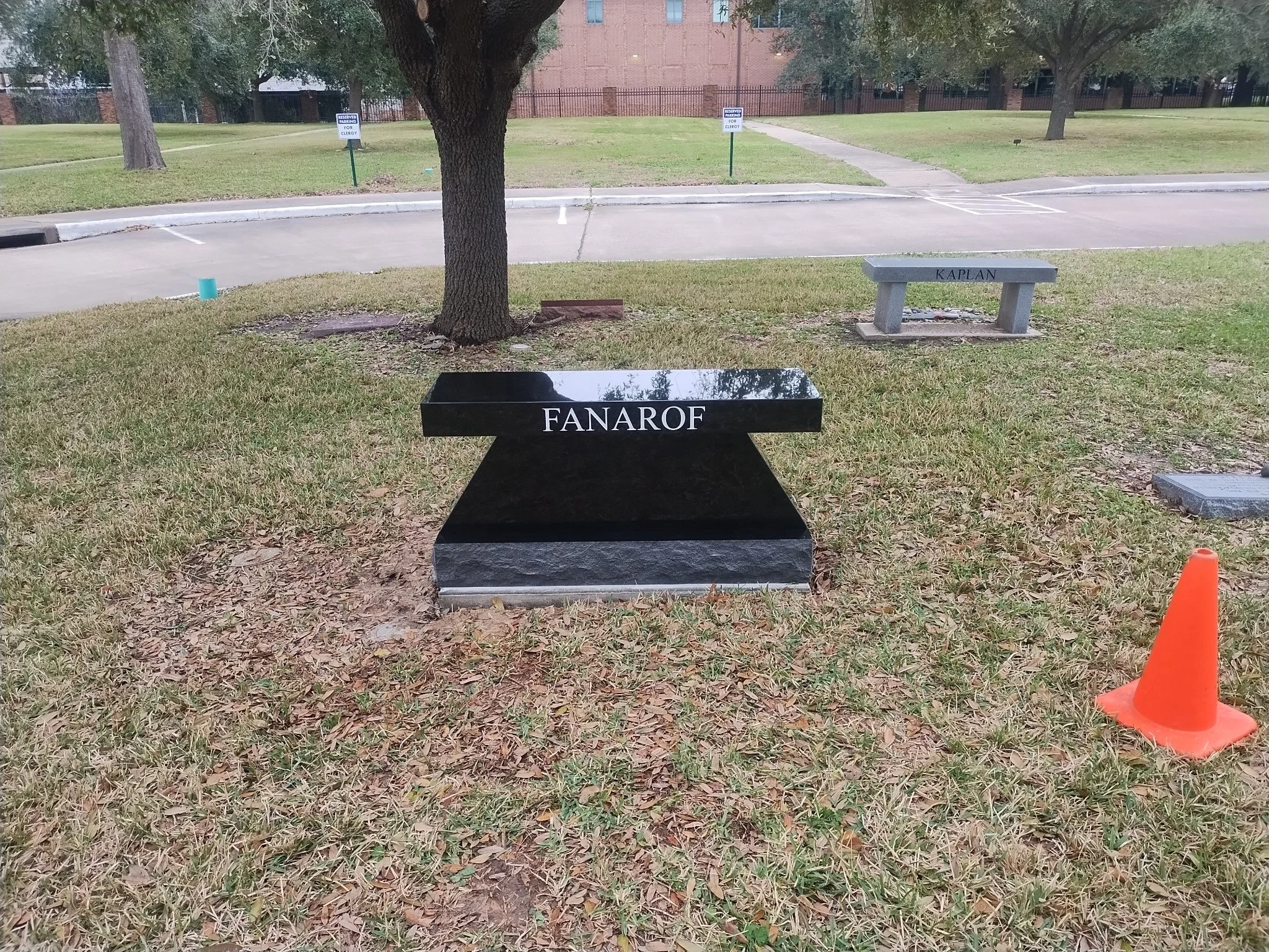 A small park with grass, trees, and a sidewalk. There are signs that say 'Reserved Parking for Clergy,' a granite bench labeled 'KAPLAN,' and a polished black stone marker labeled 'FANAROF.' An orange traffic cone is in the bottom right corner.