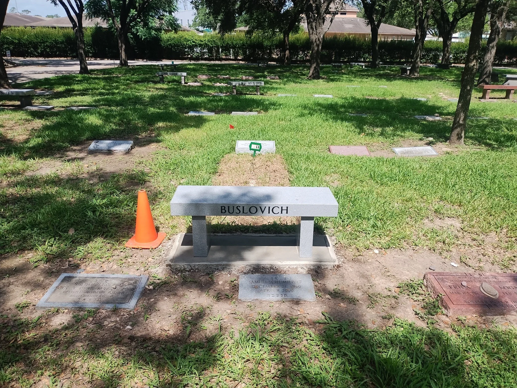 A cemetery with multiple rows of flat grave markers and lamps, under trees in a park-like setting, with a bench in the middle bearing the name 'BUSLOVICH'.