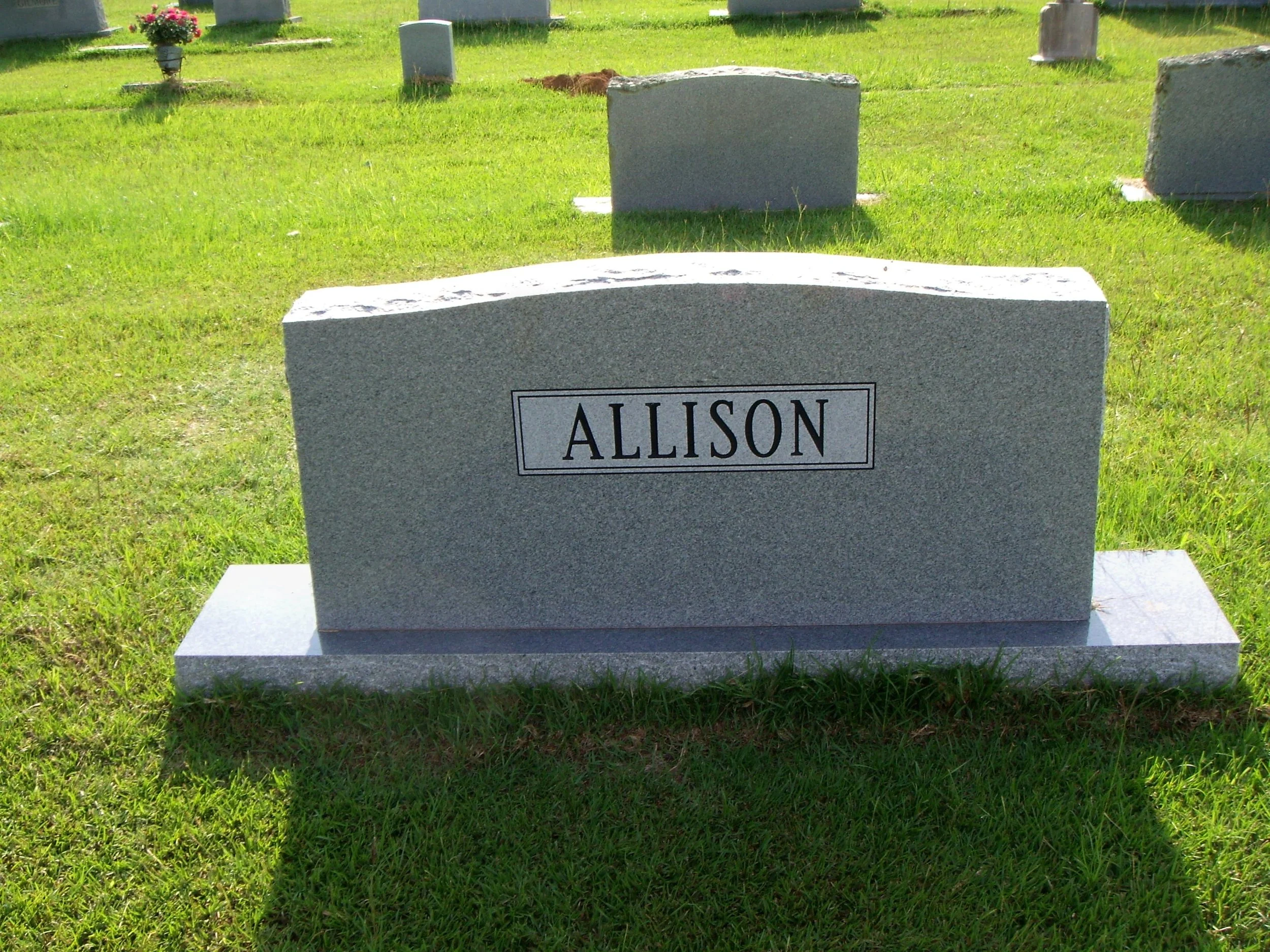 Gravestone engraved with the name in a cemetery, surrounded by other headstones and green grass.