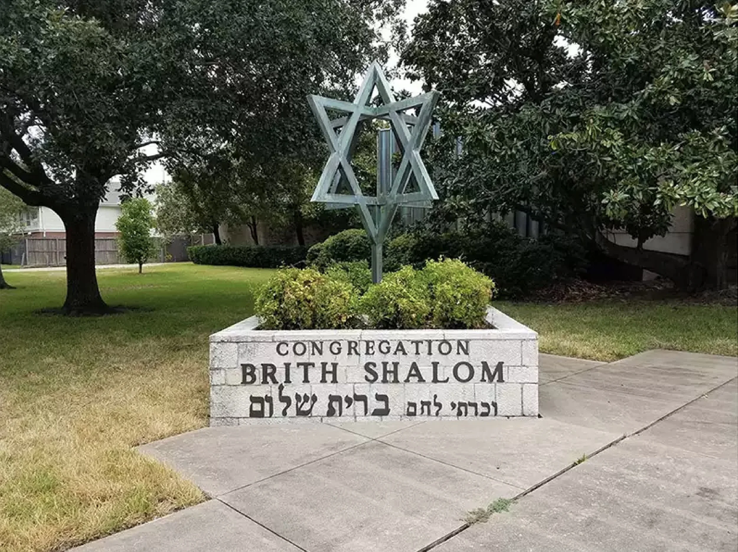 A Jewish memorial garden with a Star of David sculpture on a pedestal surrounded by green bushes and grass, with trees and buildings in the background.