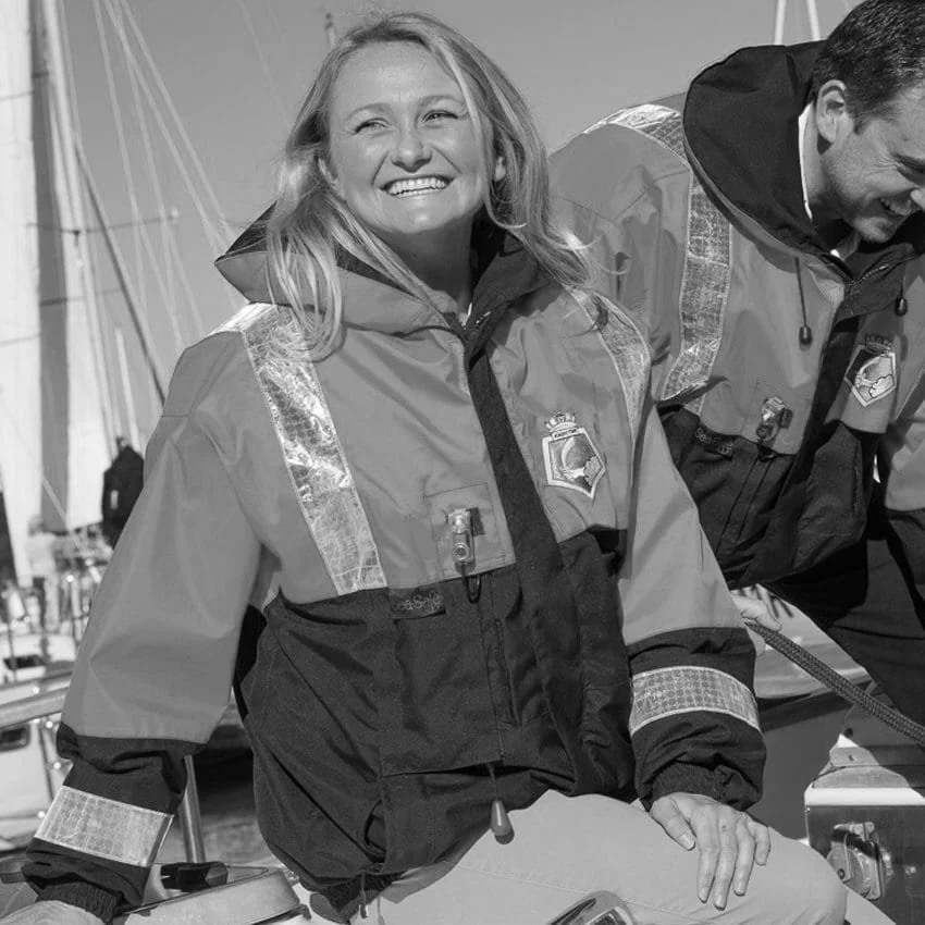 Black-and-white photo of a smiling woman wearing a maritime safety jacket aboard a vessel, with another crew member in the background.