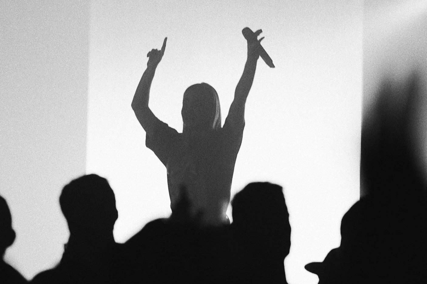 Silhouette of a woman with arms raised, holding a microphone, in front of an audience, black and white photo.