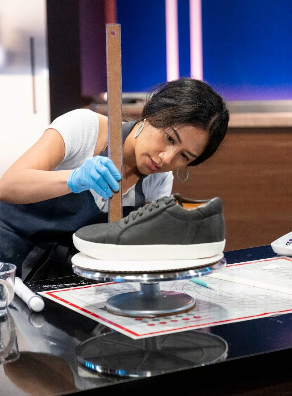 April Julian wearing a black apron and blue gloves is decorating a black sneaker on a cake stand, using a chocolate bar as a ruler. She is leaning over a table with papers and a white marker, focused on her work.