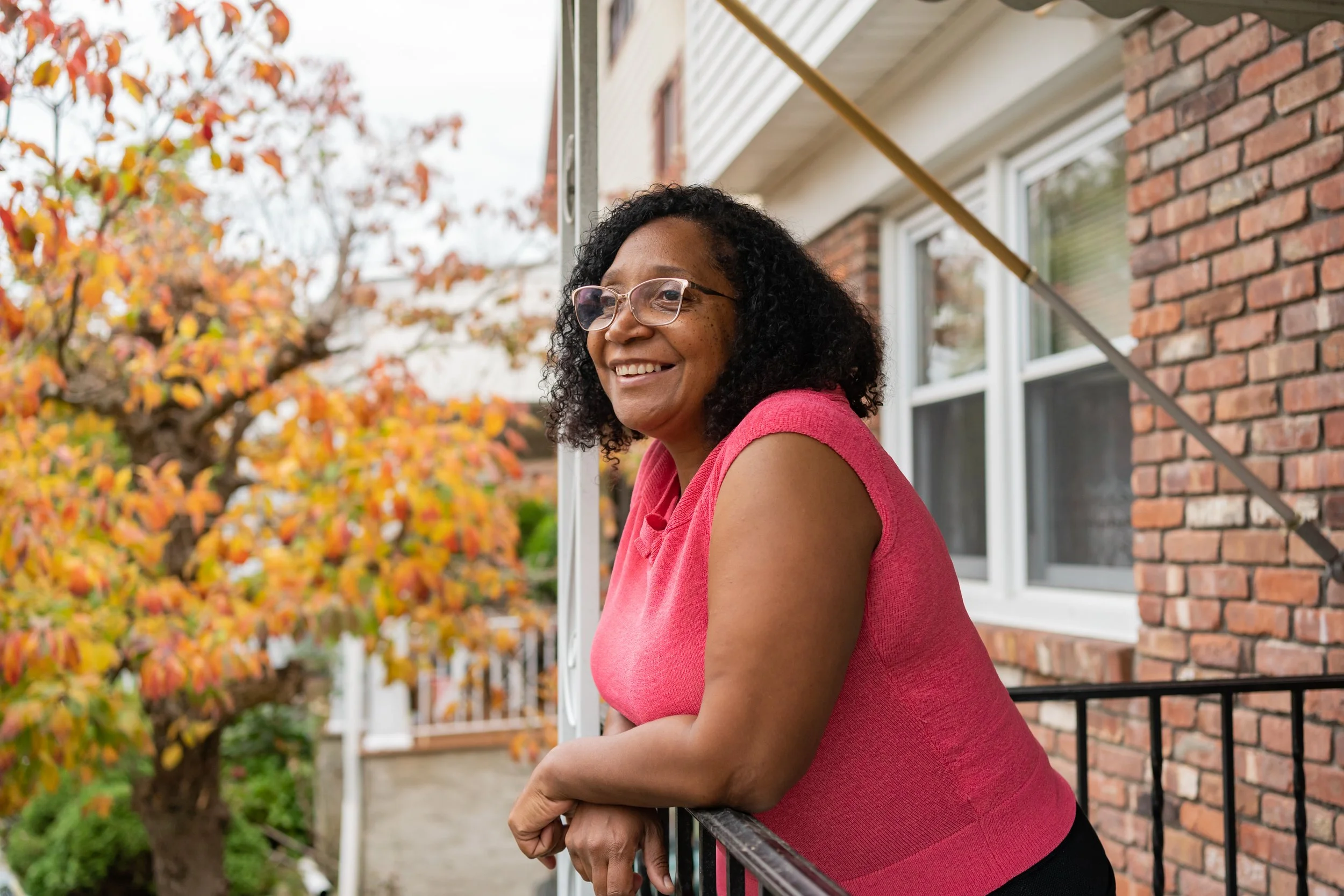 person with glasses and a pink shirt smiles and leans on a railing outside of a brick house next to fall foliage
