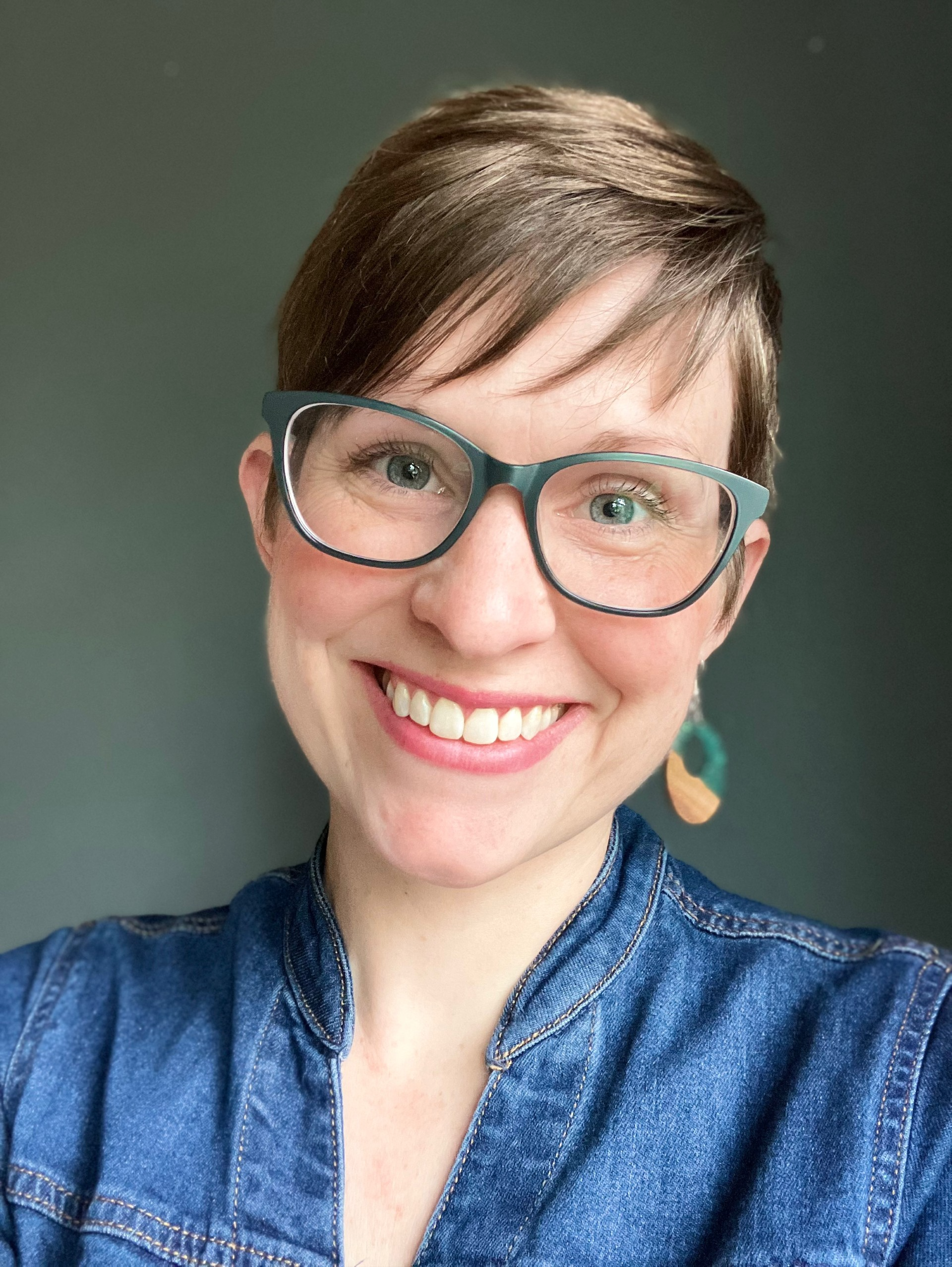 A smiling woman with short brown hair and glasses, wearing a denim jacket, with a dark plain background.