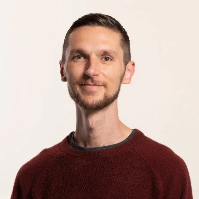 A young man with short brown hair and a beard is standing against a plain off-white background. He is wearing a maroon sweater over a dark shirt and is smiling gently at the camera.