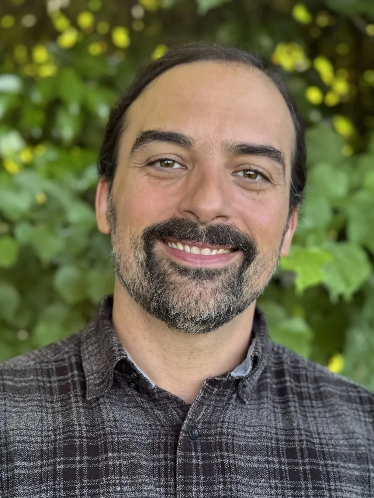 A man with dark hair and a beard smiling outdoors in front of a lush green leafy background, wearing a dark patterned shirt.