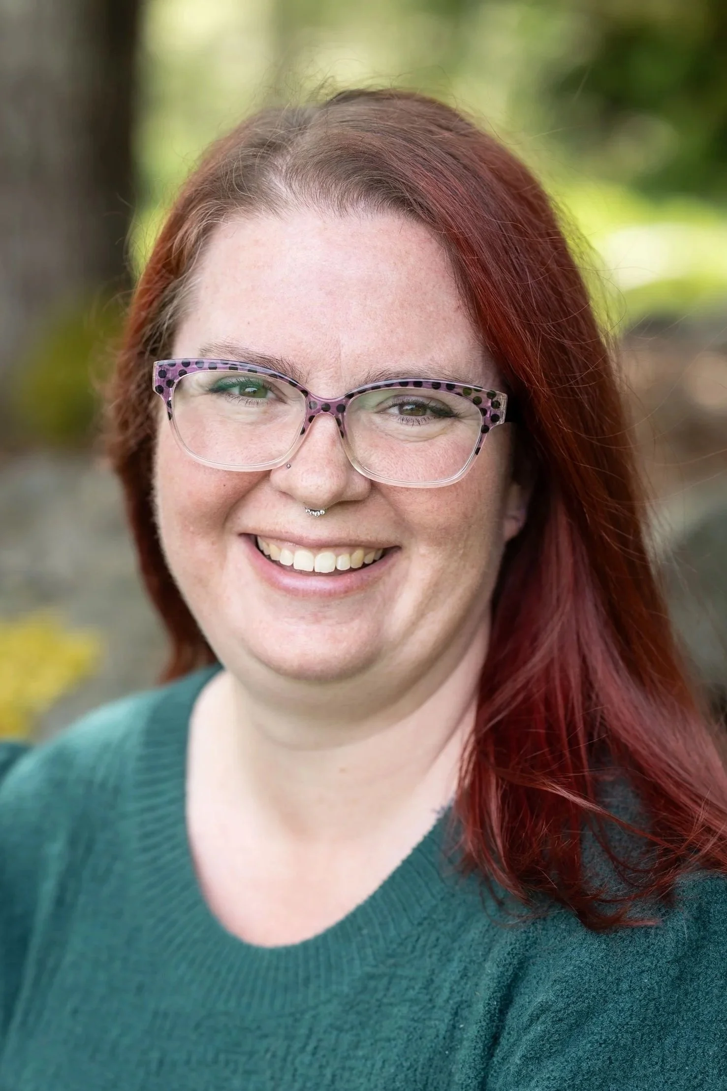 A woman with red hair, glasses, and a green sweater smiling outdoors with trees and rocks in the background.