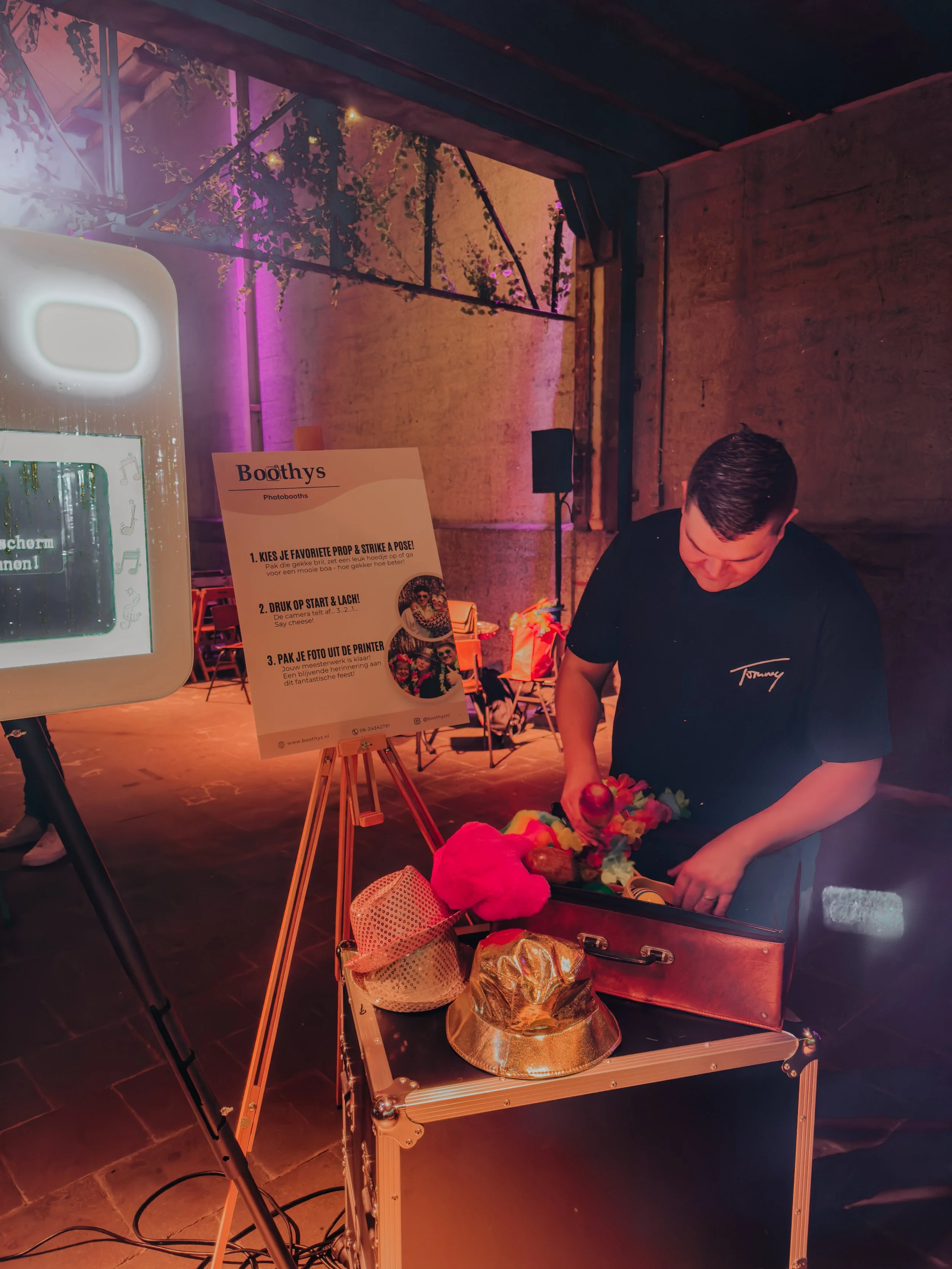 Een persoon staat bij een photobooth met een bord met instructies en verschillende accessoires zoals hoeden en een kleurrijke boa. Het is een indoor setting met sfeerverlichting.