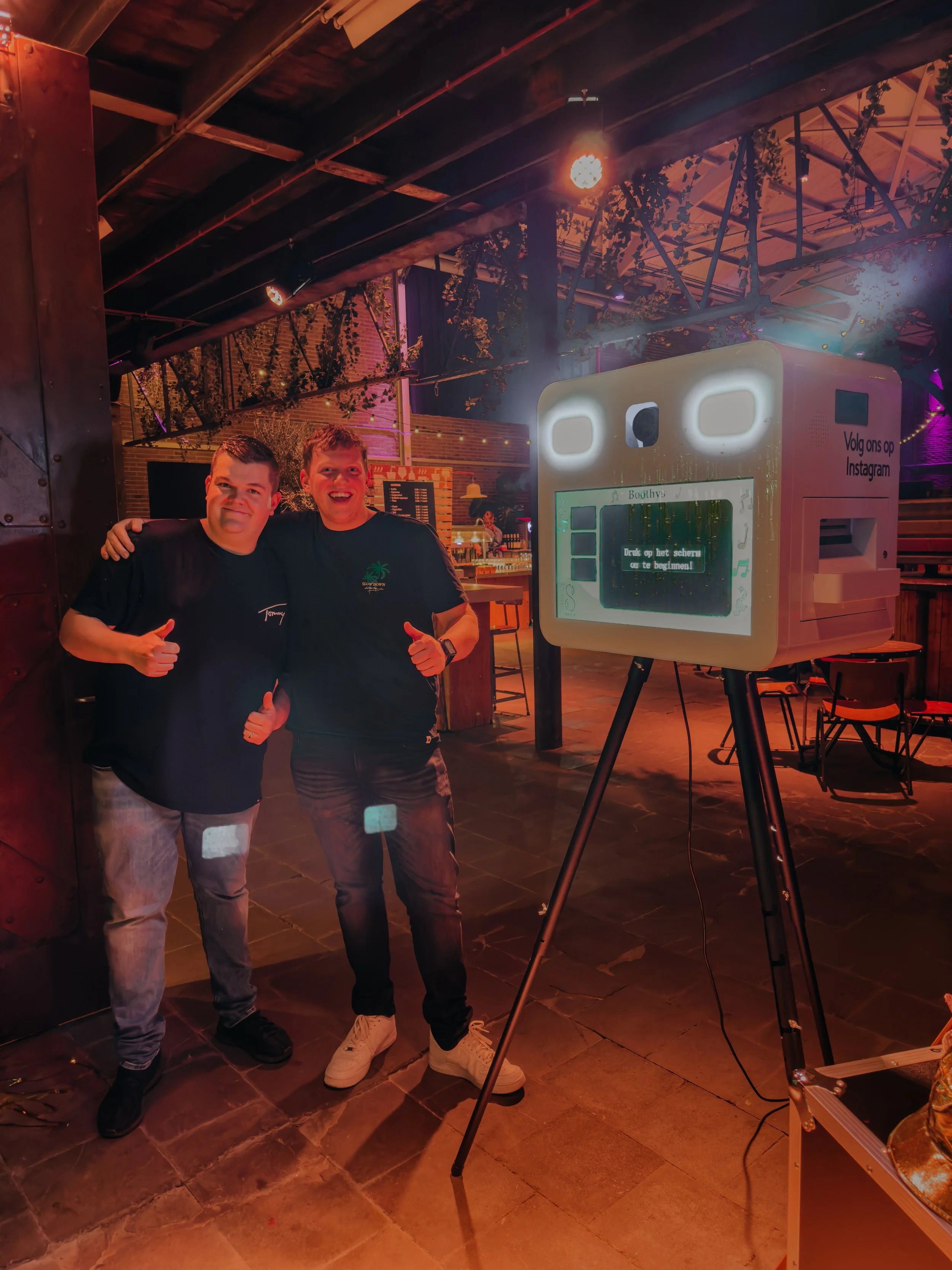 Twee mannen poseren naast een fotohokje in een feestelijke omgeving.