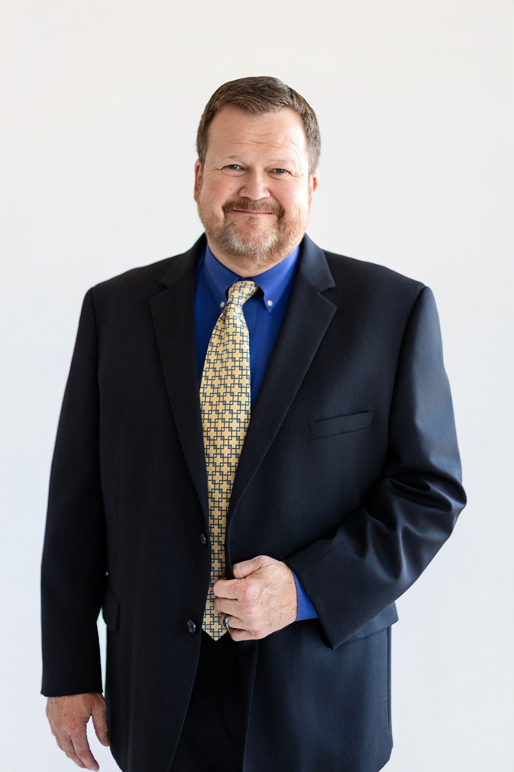 A middle-aged man with a beard, wearing a dark suit, blue shirt, and yellow patterned tie, standing against a plain white background, smiling softly.