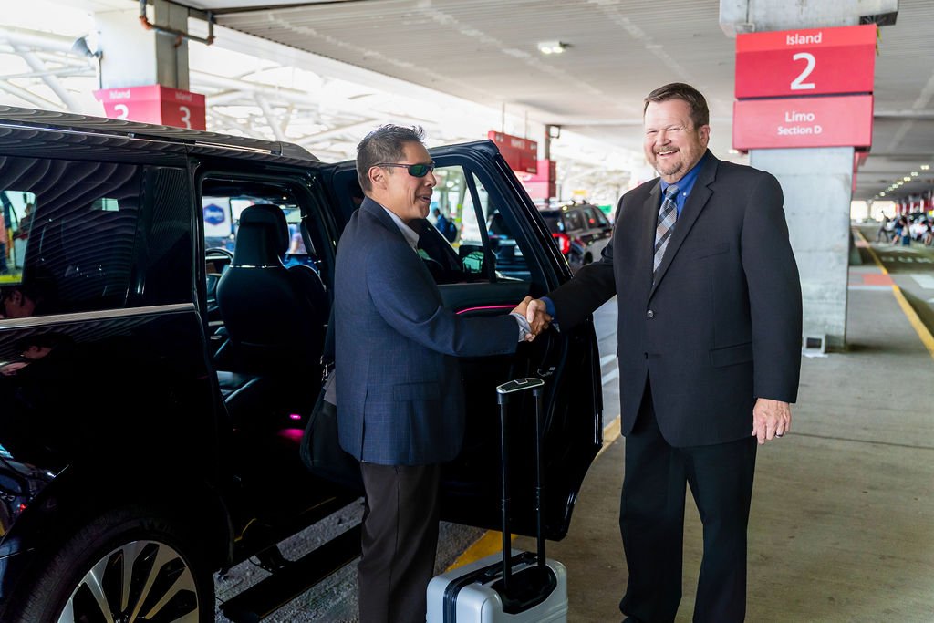 Two men in suits shaking hands at luggage pickup area of an airport, one with sunglasses and carry-on luggage, both smiling.