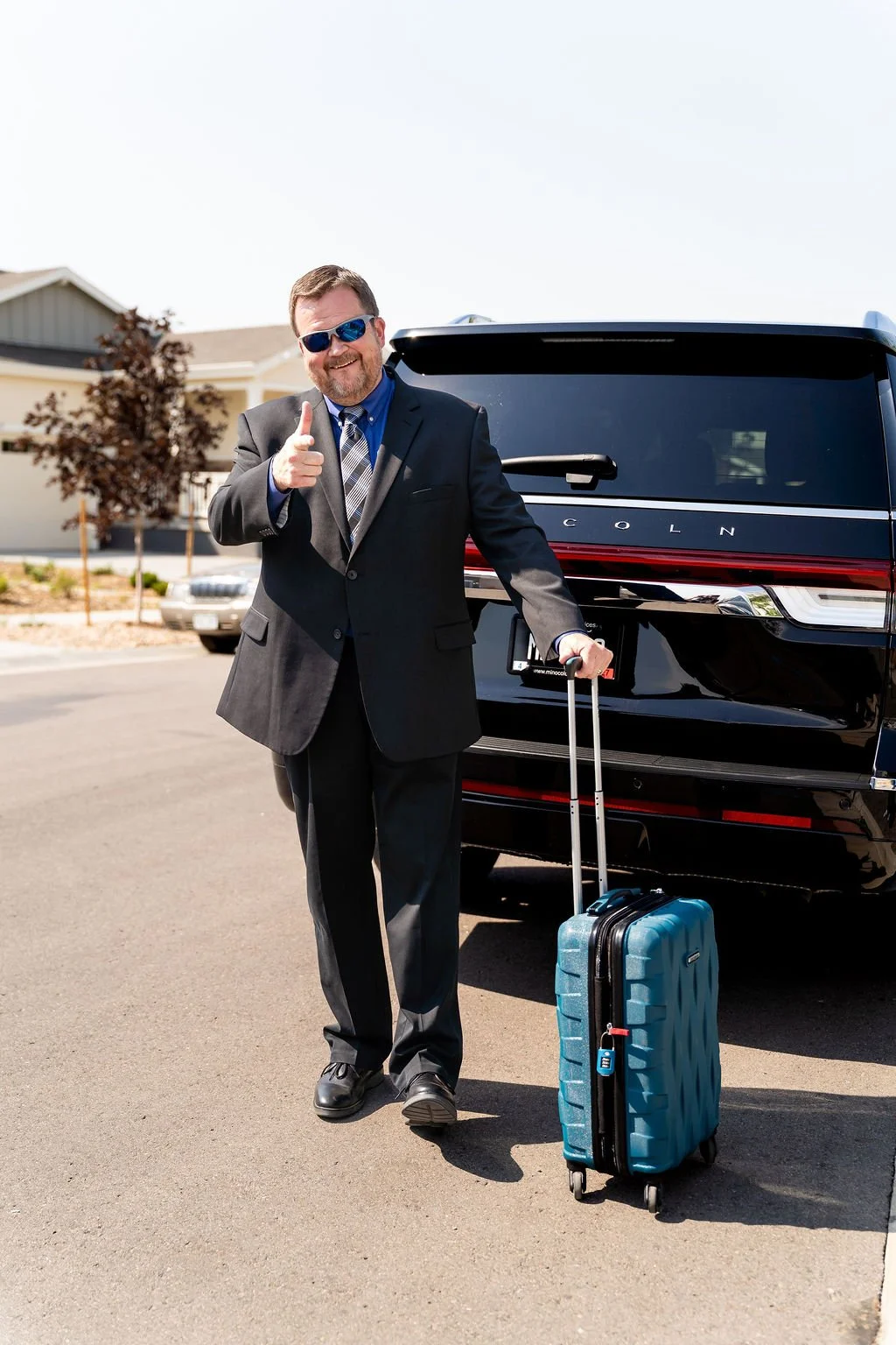 A man in a suit with sunglasses standing next to a black Lincoln SUV, holding a blue rolling suitcase, giving a thumbs up.