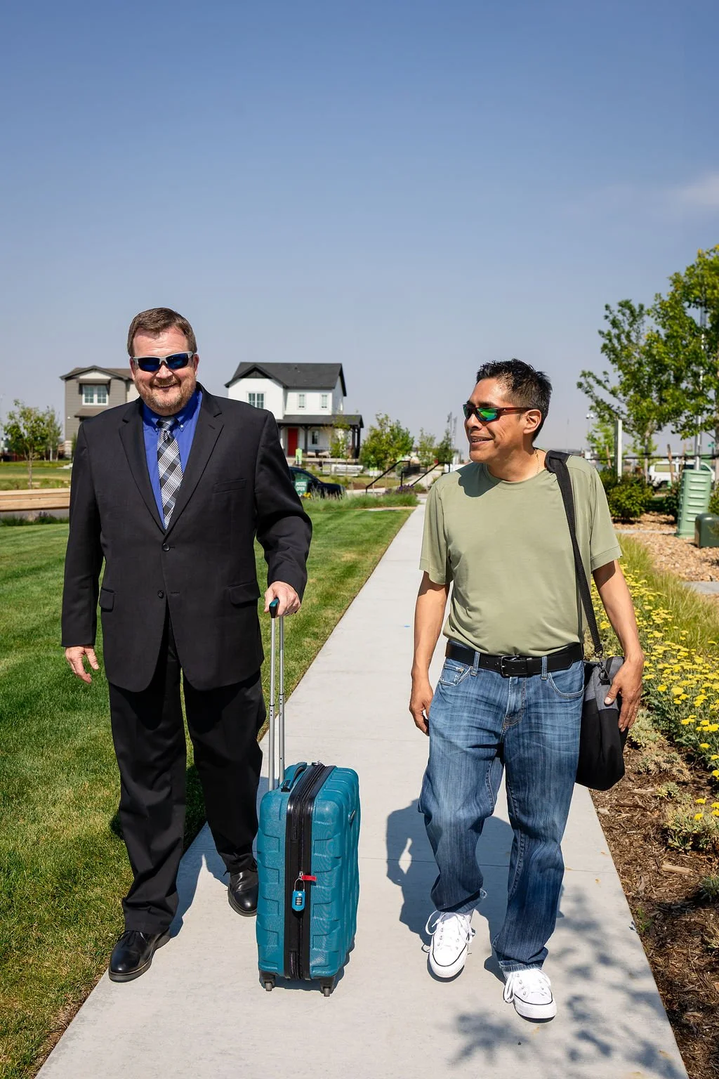 Mino Limo driver and a business man walking on a sidewalk in a residential neighborhood, one dressed in a suit with a rolling suitcase, the other in casual clothes with a bag over his shoulder, both wearing sunglasses and smiling.