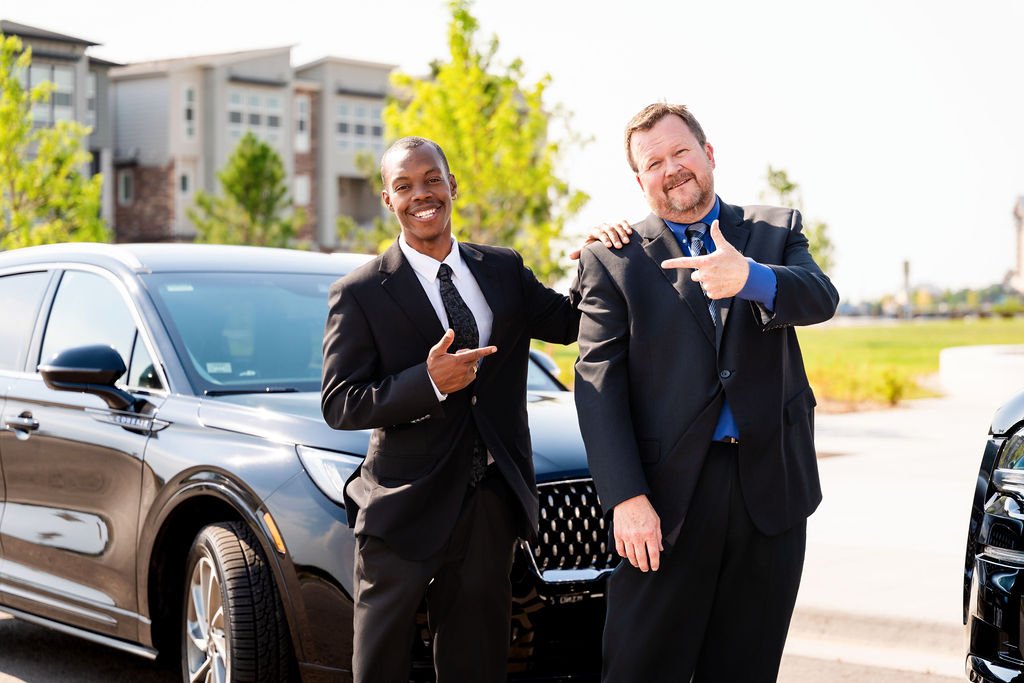 Two businessmen in suits standing outdoors next to a black sedan, smiling and pointing at the car, with modern buildings and trees in the background.