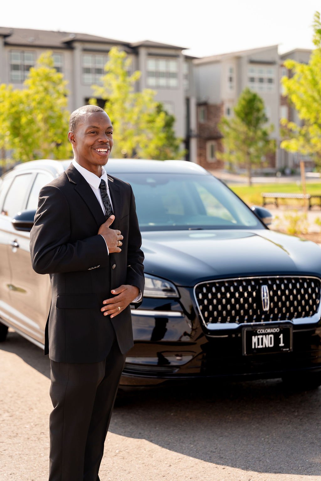 A young man in a suit standing next to a black Lincoln car with a Colorado license plate that reads 'MINO 1,' smiling and placing his right hand on his chest.