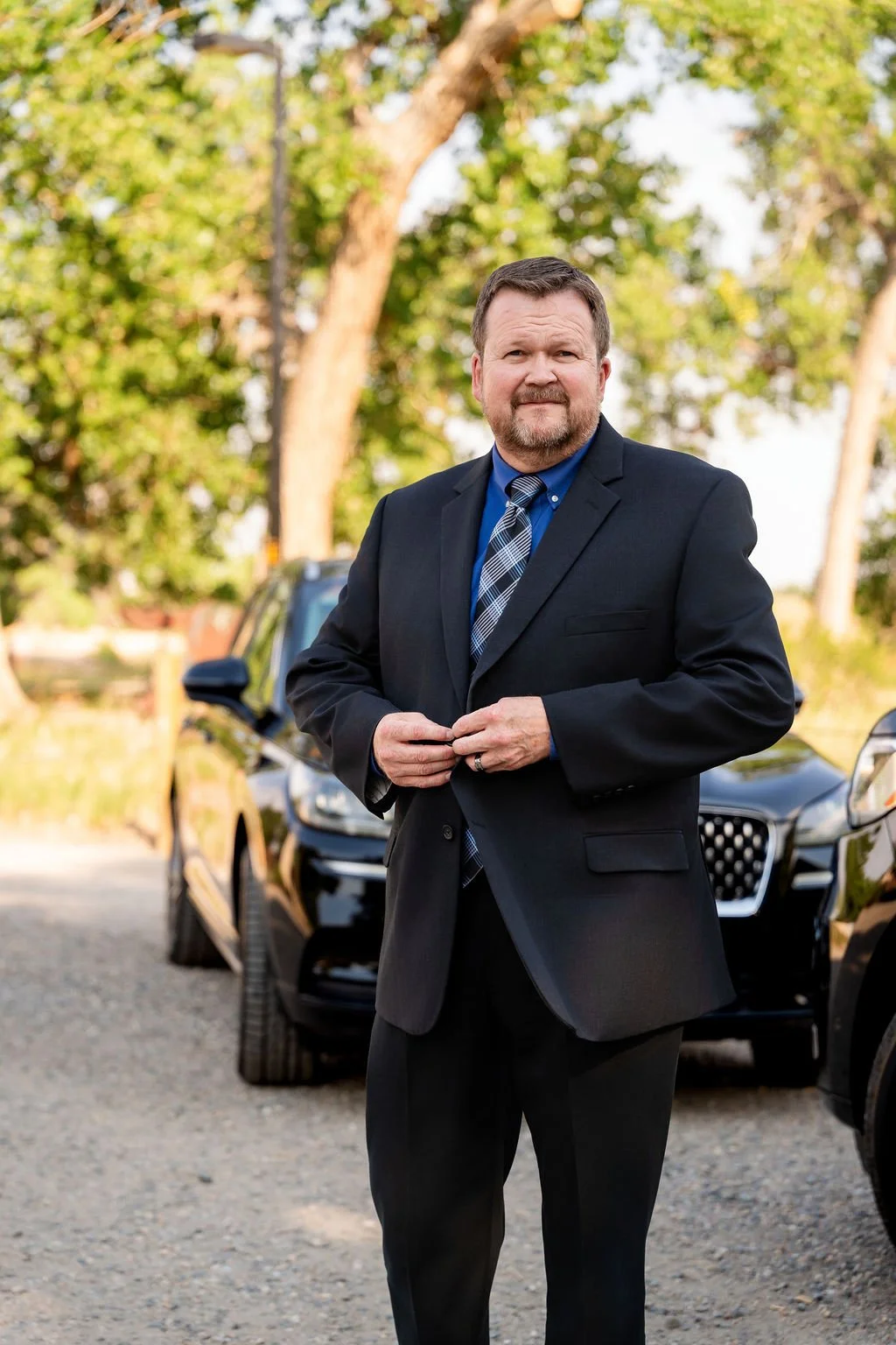 A man in a suit standing outdoors in front of cars and trees.