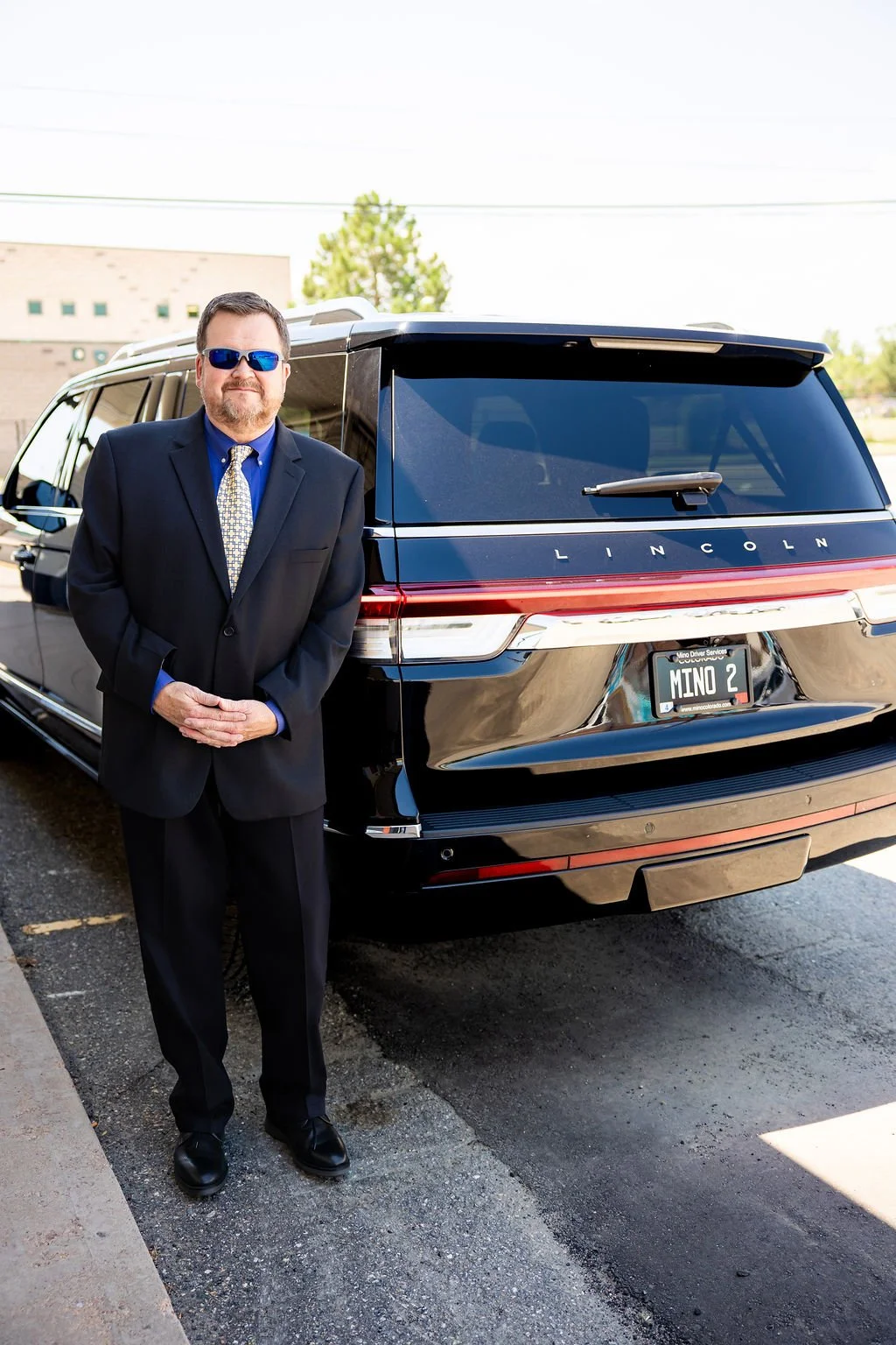 Limo driver in a black suit and a patterned tie, wearing blue sunglasses, standing beside a black Lincoln SUV with a custom license plate reading 'MINO 2'.