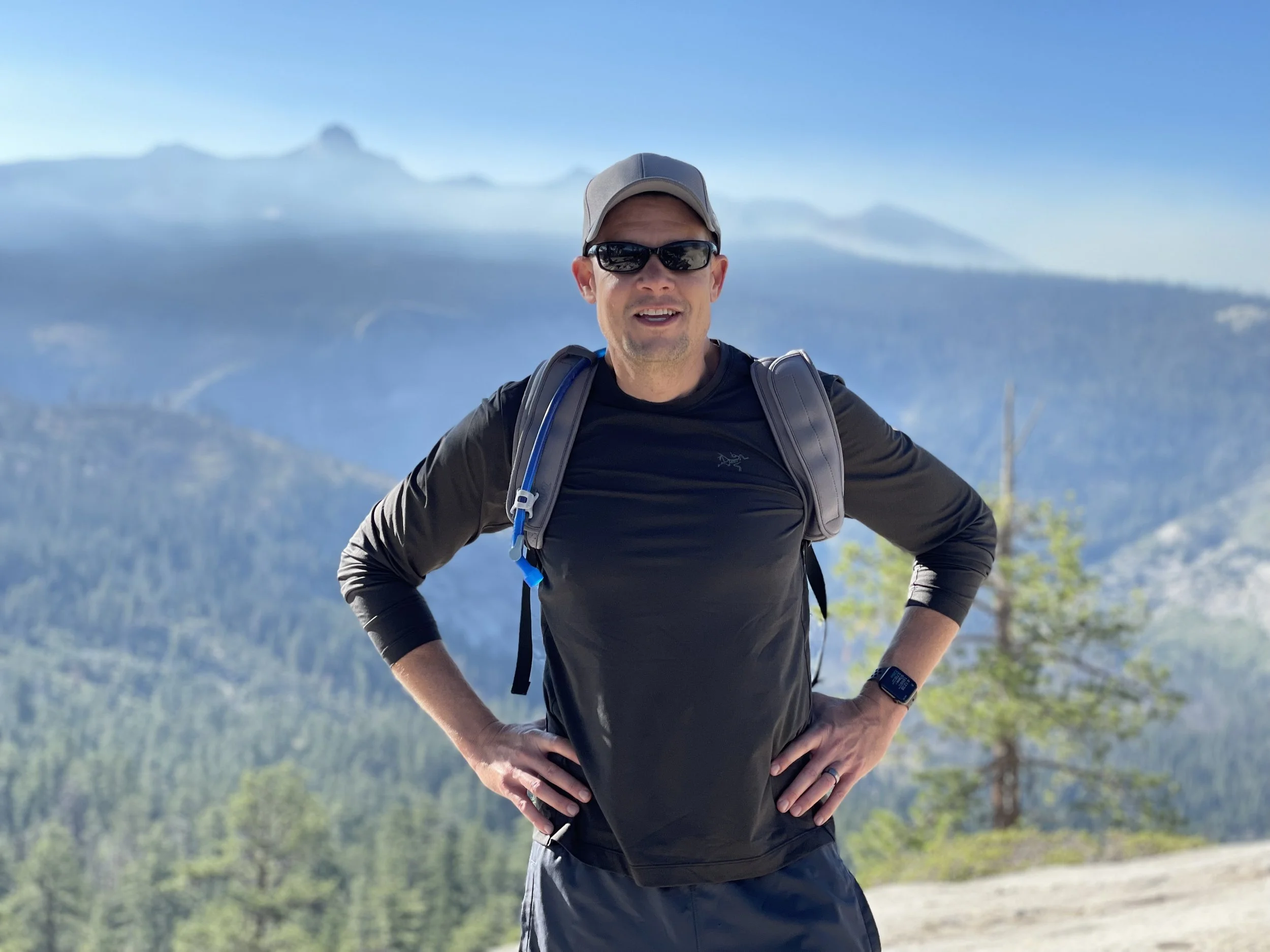 A man wearing a gray cap, black sunglasses, and a black long-sleeve shirt standing outdoors on a trail with mountain and forest landscape in the background. He has a backpack and is smiling with his hands on his hips.