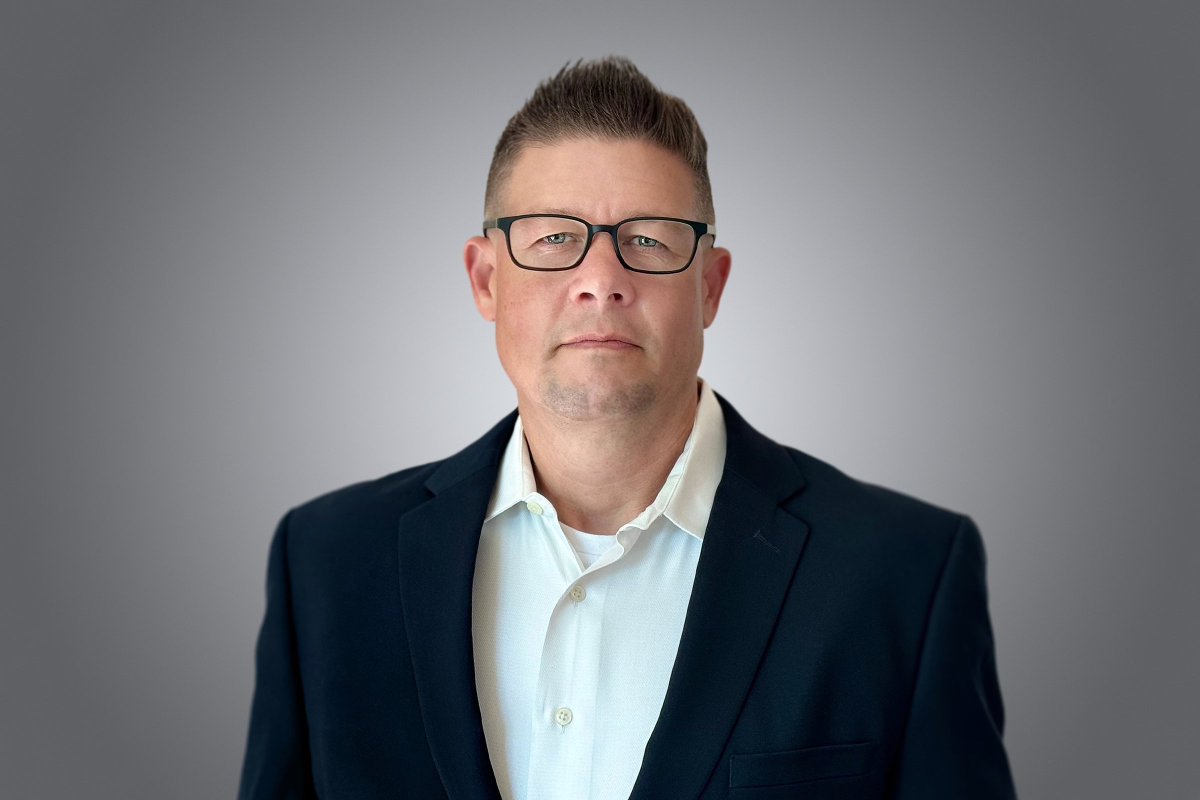 A man with glasses, short brown hair, wearing a white collared shirt and a dark suit jacket, standing against a plain gray background.