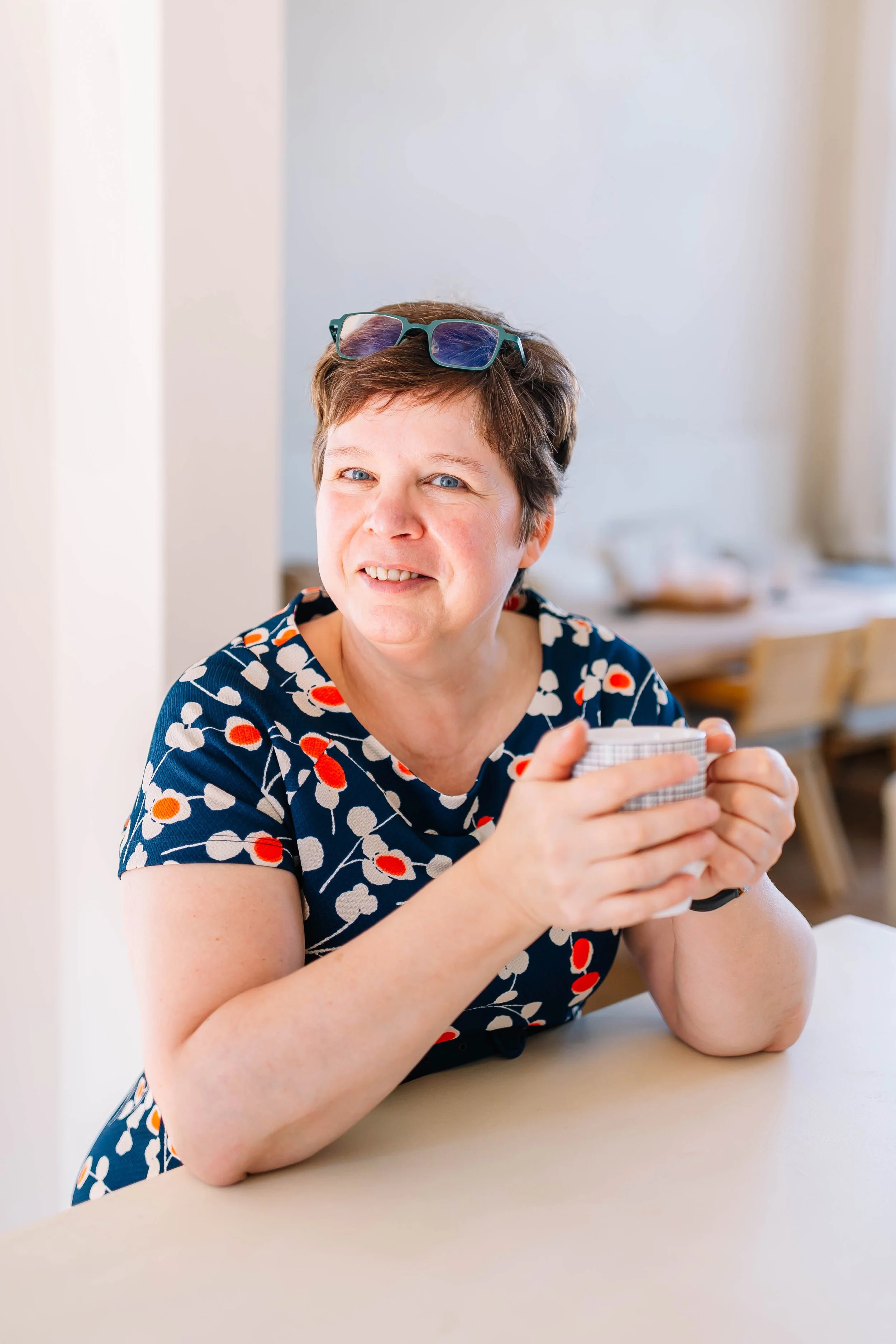 Hilde - oprichtster van 'BRAAZH': Een vrouw met kort bruin haar, blauwe ogen en een speelse glimlach zit aan een tafel, houdt een mok met beide handen vast, draagt een blouse met een bloemmotief, en heeft een paar zonnebrillen op haar hoofd.